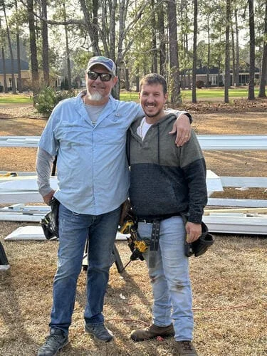 Two crew members in tool belts standing beside white railing and trim pieces laid out on pine straw outdoors