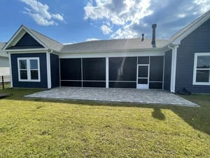 Dark screen enclosure with white trim and gray paver patio behind a navy blue home with white-trimmed windows